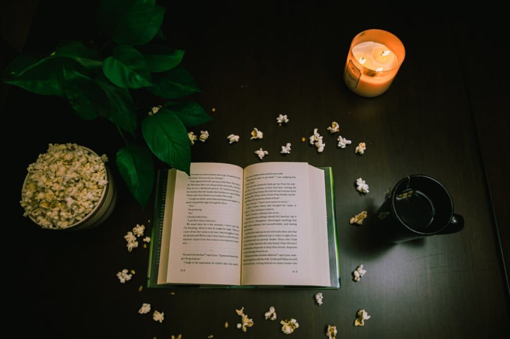 An open book surrounded by scattered popcorn, a candle, and a mug on a dark surface