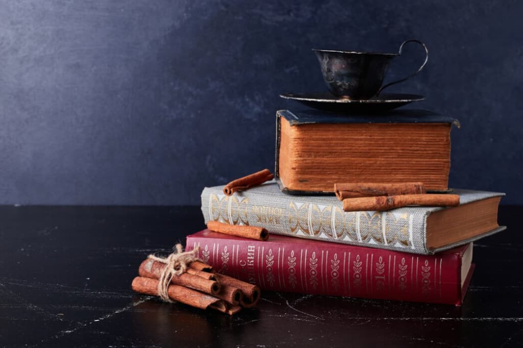 A vintage cup on top of a pile of old books with cinnamon sticks on a dark table