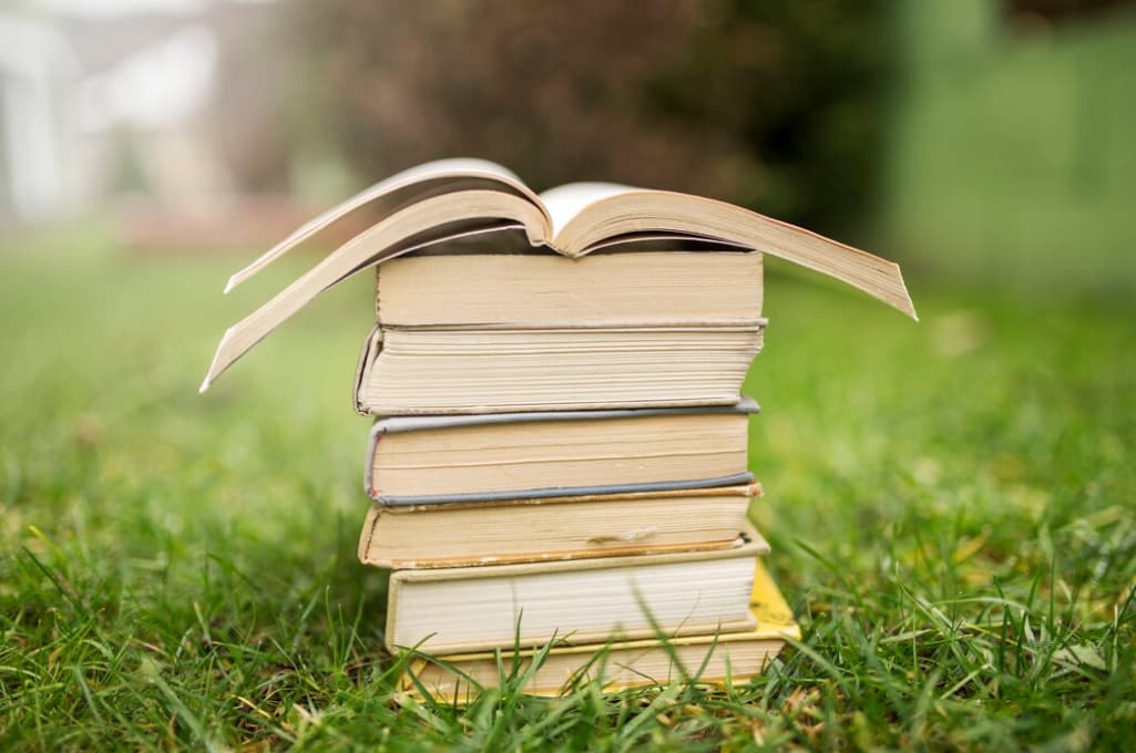 A stack of hardcover books with an open one on top, set on a grassy lawn with a blurred background