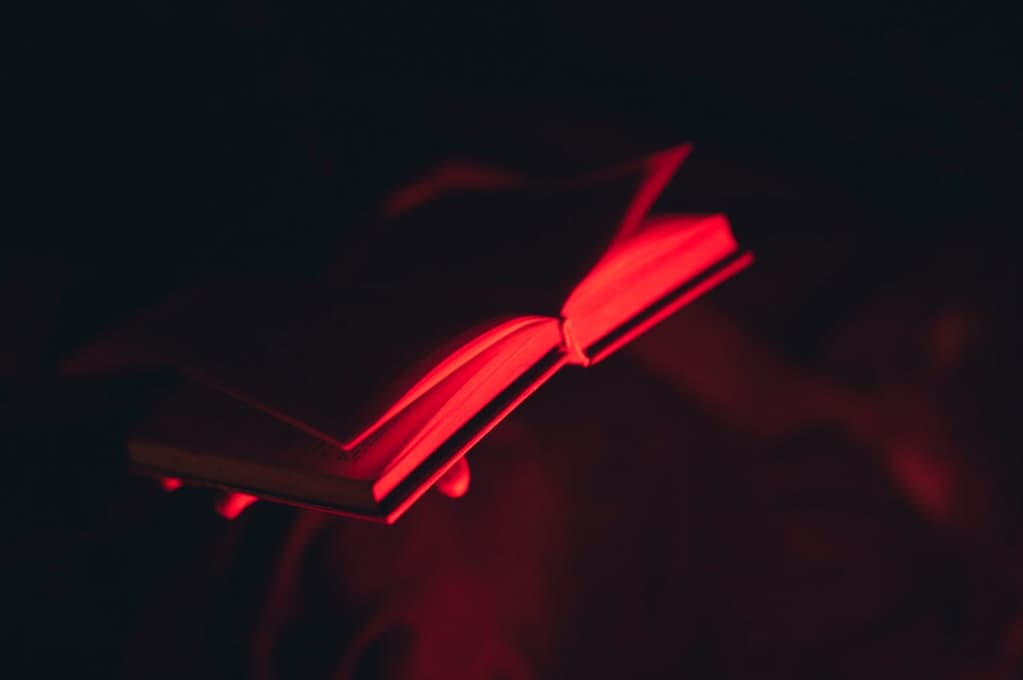 A glowing red light illuminates an open book in a dark, moody setting